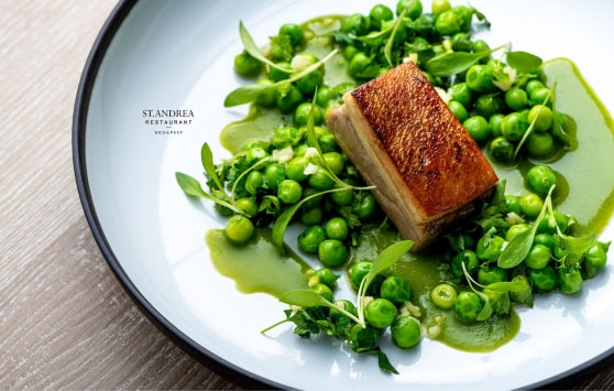 Roast duck with green peas, leafy greens and green sauce served on a white plate.